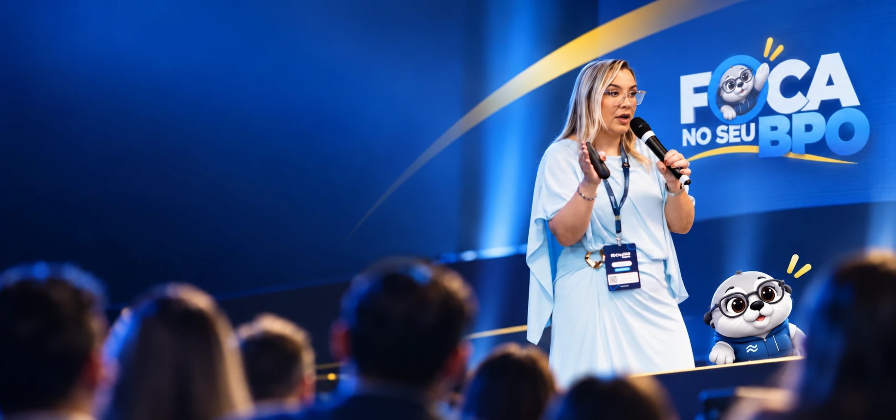Cláudia Bernardo no palco do Foca no seu BPO, falando para a plateia, com a logo do evento e a mascote foca ao fundo